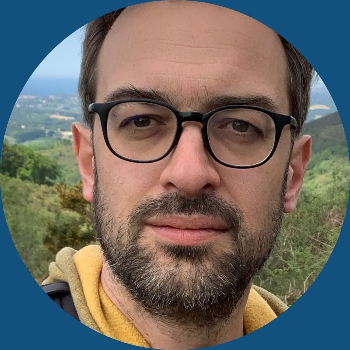 Image of Dr Nicolas Fortane, a man with pale-skin, short dark har, a short dark beard and glasses. He is wearing a yellow hooded shirt and is standing in front of an outdoor view.